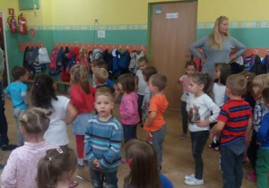 Dzieci stojące w sali przedszkolnej naśladują ruchy nauczycielek – trzymają ręce pod boki.